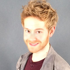 Redheaded Michael Geiser wearing a grey jacket over a maroon shirt in front of a grey background