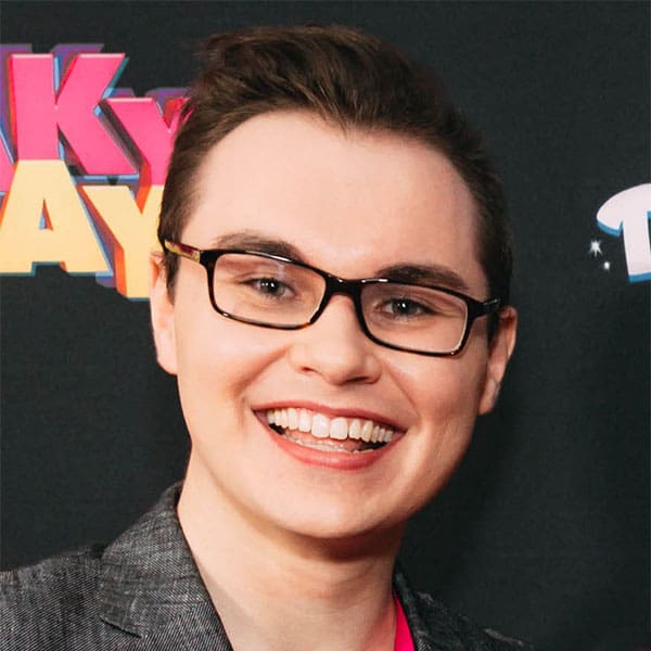 Stephen Fertelmes in black glasses and a checked grey sportscoat in front of a black background with pink and yellow letters on it