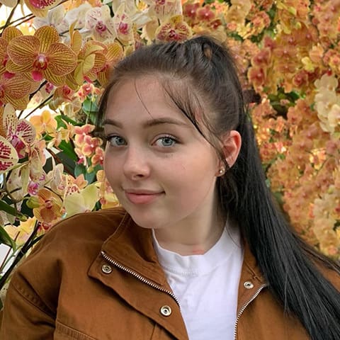 Kyla Chalmers in a brown jacket in front of a bunch of yellow and orange flowers, looking quizzically into the camera