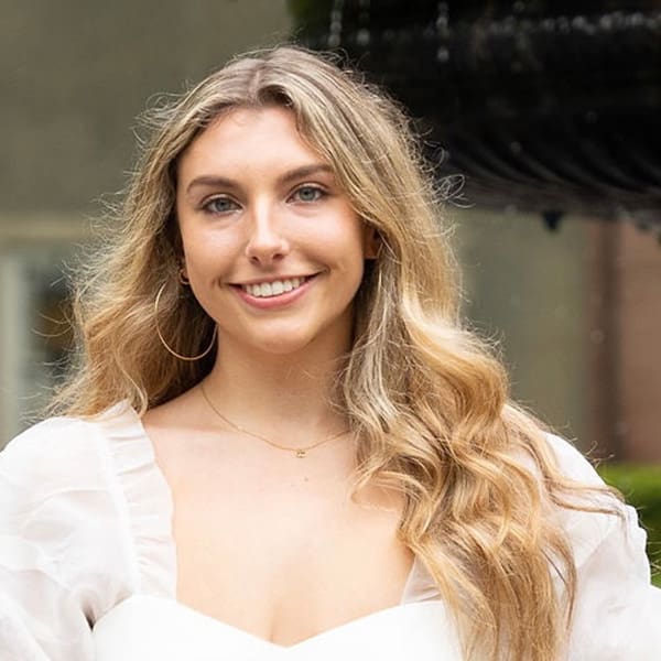 Chloe Cockery in a white dress in front of a fountain, smiling into the camera