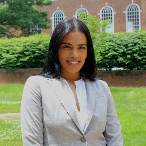 Rachel Bencosme in a gray suit jacket in front of a red brick building, smiling into the camera